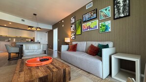 A modern living room with a grey sofa, a wooden coffee table, and a TV stand. There is a kitchen area with a white countertop and a hanging light fixture.