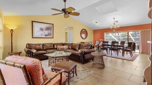 A living room with a brown leather sofa, a glass coffee table, wicker chairs, a ceiling fan, and a dining area with wooden chairs and a table. - Bokeelia Bliss! 3/3 Heated Pool, Boat Ramp Included! (Bokeelia)