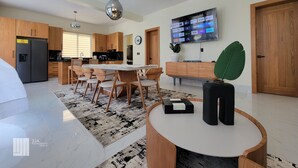 A modern kitchen with wooden cabinets, a dining area with a round table, and a TV mounted on the wall displaying various streaming services. - Villa Marina (Sosúa)