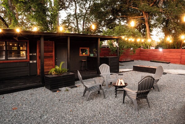 A rustic outdoor seating area with Adirondack chairs and a small fire pit, surrounded by a gravel patio and a wooden deck.