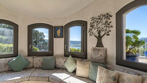 A cozy, curved sectional sofa with patterned cushions, large windows with dark frames, and a decorative tree sculpture. - Villa Sallena by Interhome (Porto Vecchio)
