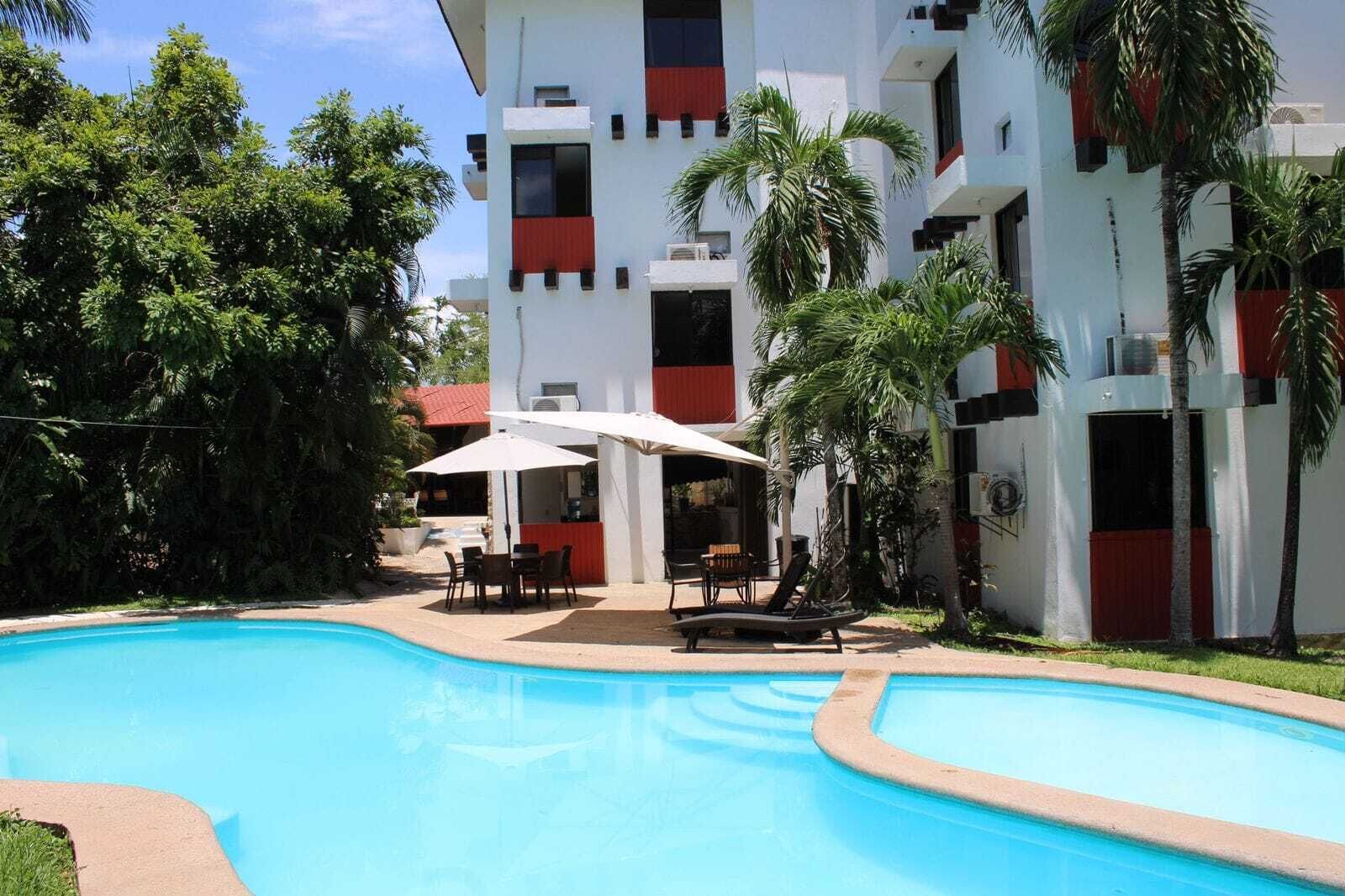 Ein Swimmingpool mit Liegestühlen und Sonnenschirmen, umgeben von Palmen und einem Gebäude mit roten Akzenten.