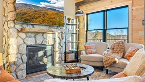 A cozy living room with a stone fireplace, a comfortable sofa, and a large window offering a scenic view. - Luxury Mountain Lodge | Hot Tub, Mtn Views, Sauna,  Game Room, Fire Pit (Bethel)
