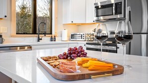 A modern kitchen with white cabinets, stainless steel appliances, a marble countertop, and a wooden board with snacks and wine glasses. - Luxury Mountain Lodge | Hot Tub, Mtn Views, Sauna,  Game Room, Fire Pit (Bethel)