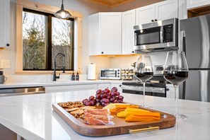Una cocina moderna con armarios blancos, electrodomésticos de acero inoxidable, encimera de mármol y una tabla de madera con aperitivos y copas de vino.