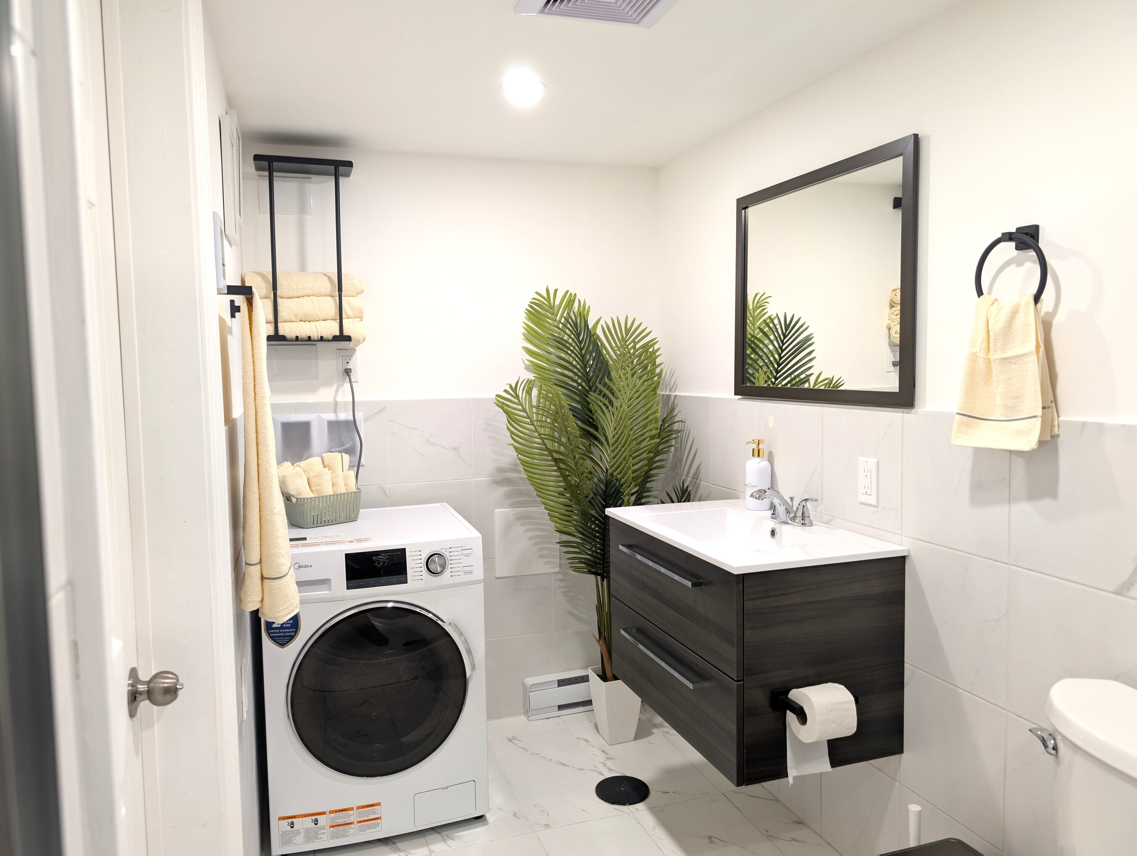 Un baño moderno con lavadora y secadora integradas, un espejo grande, un lavamanos con toallero y una planta en maceta.