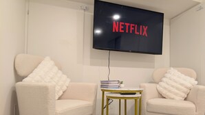 Una sala de estar moderna con dos sillones cómodos, una mesita de noche con libros y un televisor en la pared mostrando “NETFLIX”.