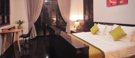 A hotel room with a bed, a desk, a chair, a potted plant, and a view of the outside.