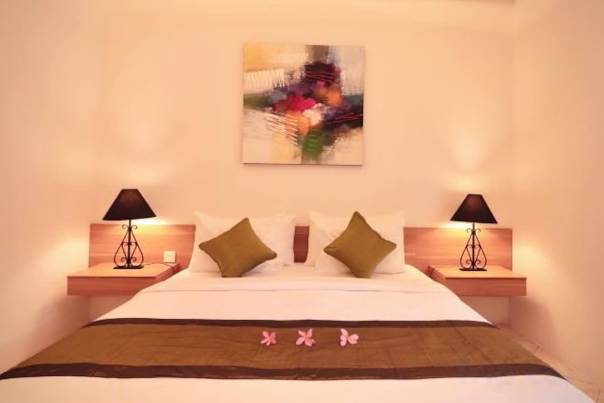 A hotel room with a bed, two bedside lamps, a wall-mounted abstract painting, and a decorative pillow arrangement.
