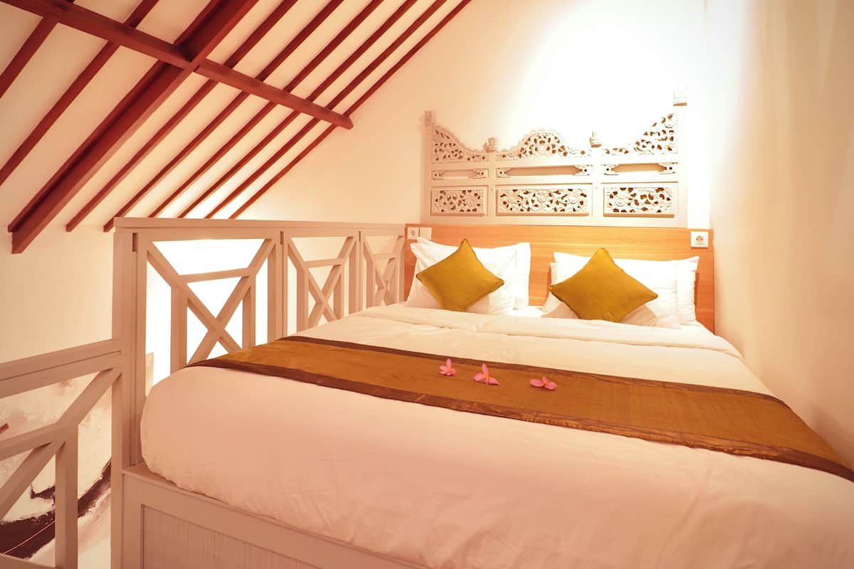 Ein Schlafzimmer mit schräg zulaufender Decke, einem hölzernen Kopfteil, einem Bett mit weißer Bettwäsche und zwei Kissen sowie einer dekorativen Wandverkleidung über dem Bett.
