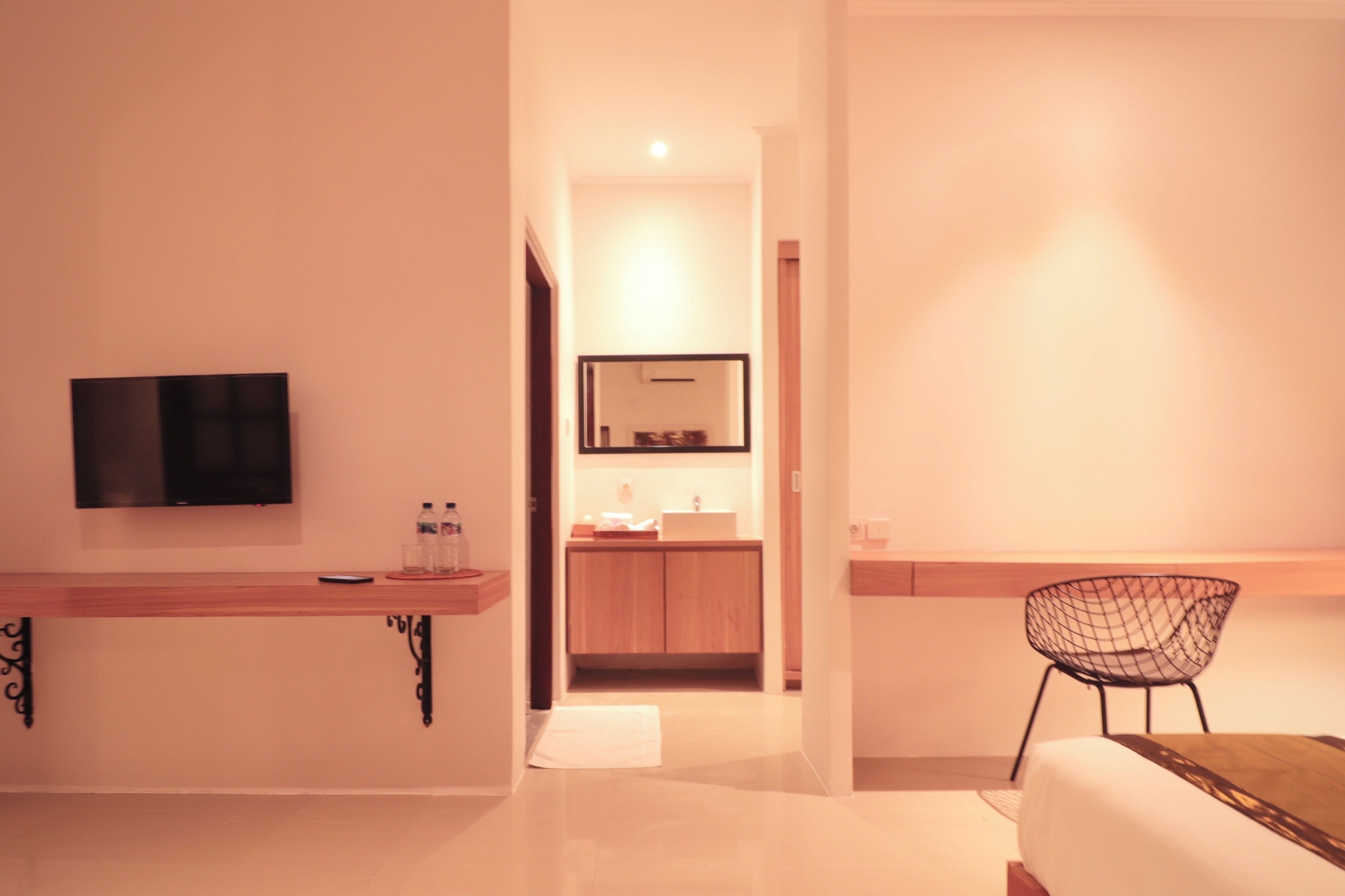 A modern hotel room with a wooden vanity, a flat-screen TV, a small sink, and a wire chair.