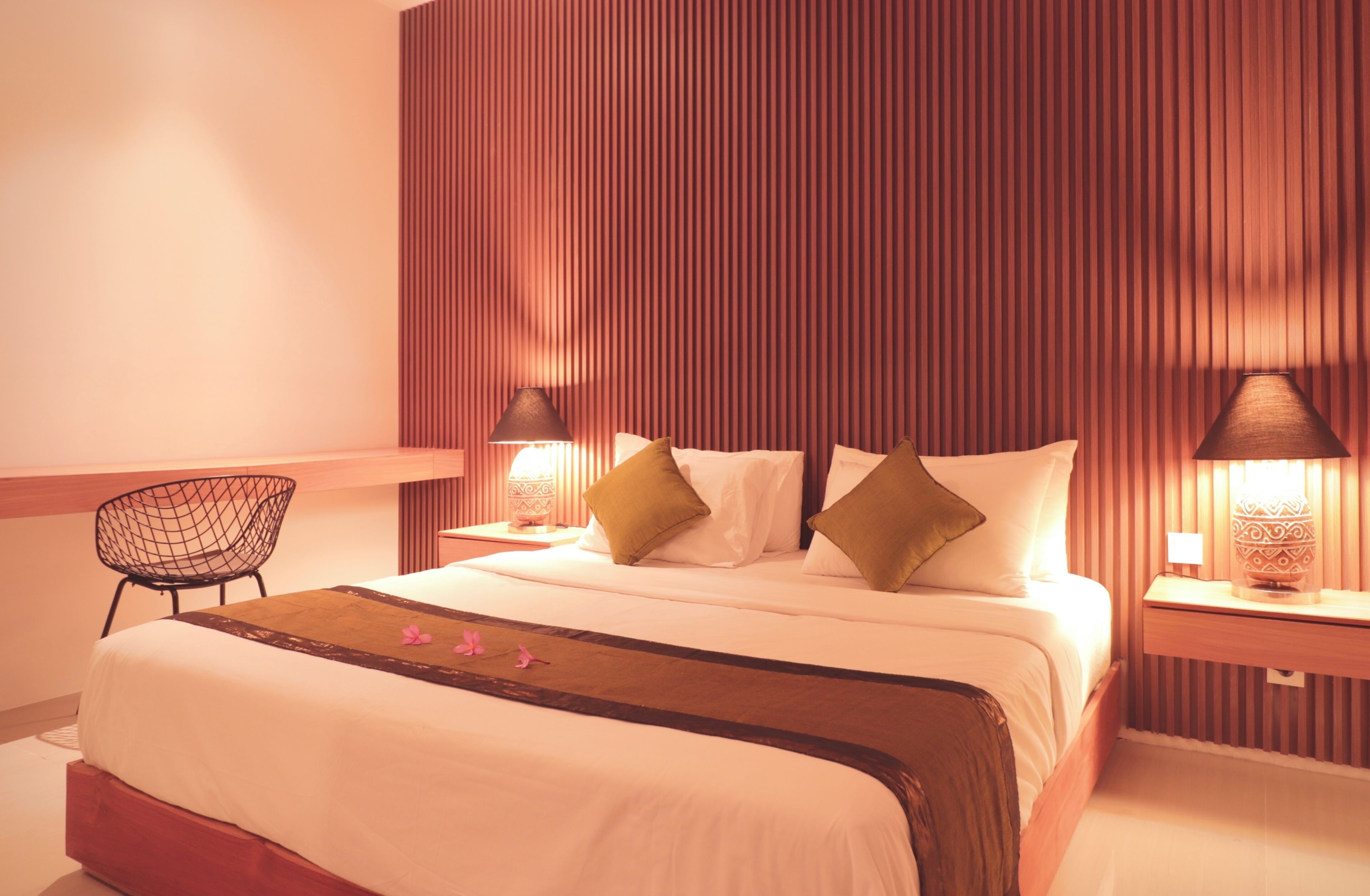 Una habitación de hotel ordenada con una cama grande, dos mesitas de noche con lámparas, una silla y un cabecero de madera.