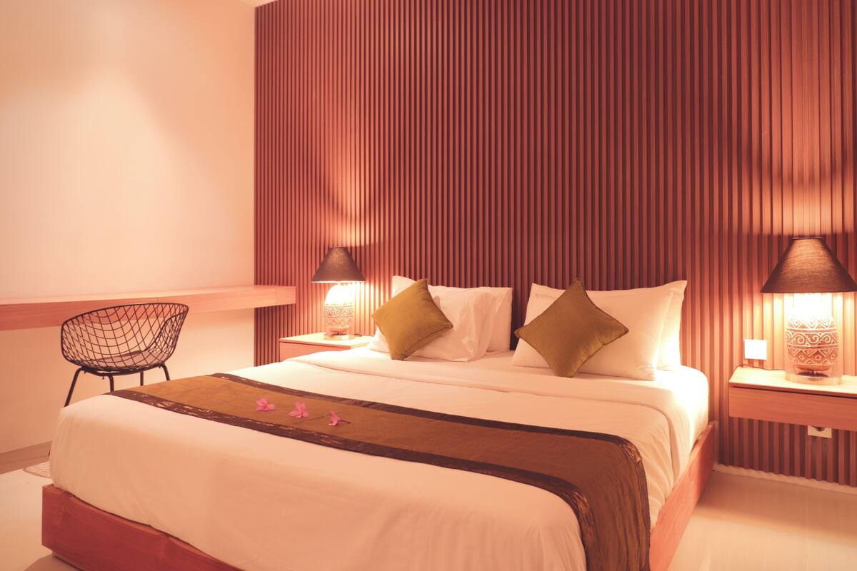 Ein ordentlich eingerichtetes Hotelzimmer mit einem großen Bett, zwei Nachttischen inklusive Lampen, einem Sessel und einem hölzernen Kopfteil.
