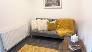 A cozy living room with a grey sofa, fluffy white and mustard throw pillows, a mustard throw blanket, a framed cityscape picture, a radiator, a yellow rug, and a small wooden side table with a dice and a book. - 1 Bed Flat - Sleeps 3 - Parking - Pet friendly (South Shields)