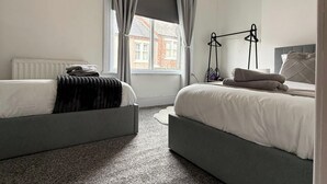 A bedroom with two beds, a radiator, a window with curtains, and a bedside table with a lamp. - 1 Bed Flat - Sleeps 3 - Parking - Pet friendly (South Shields)