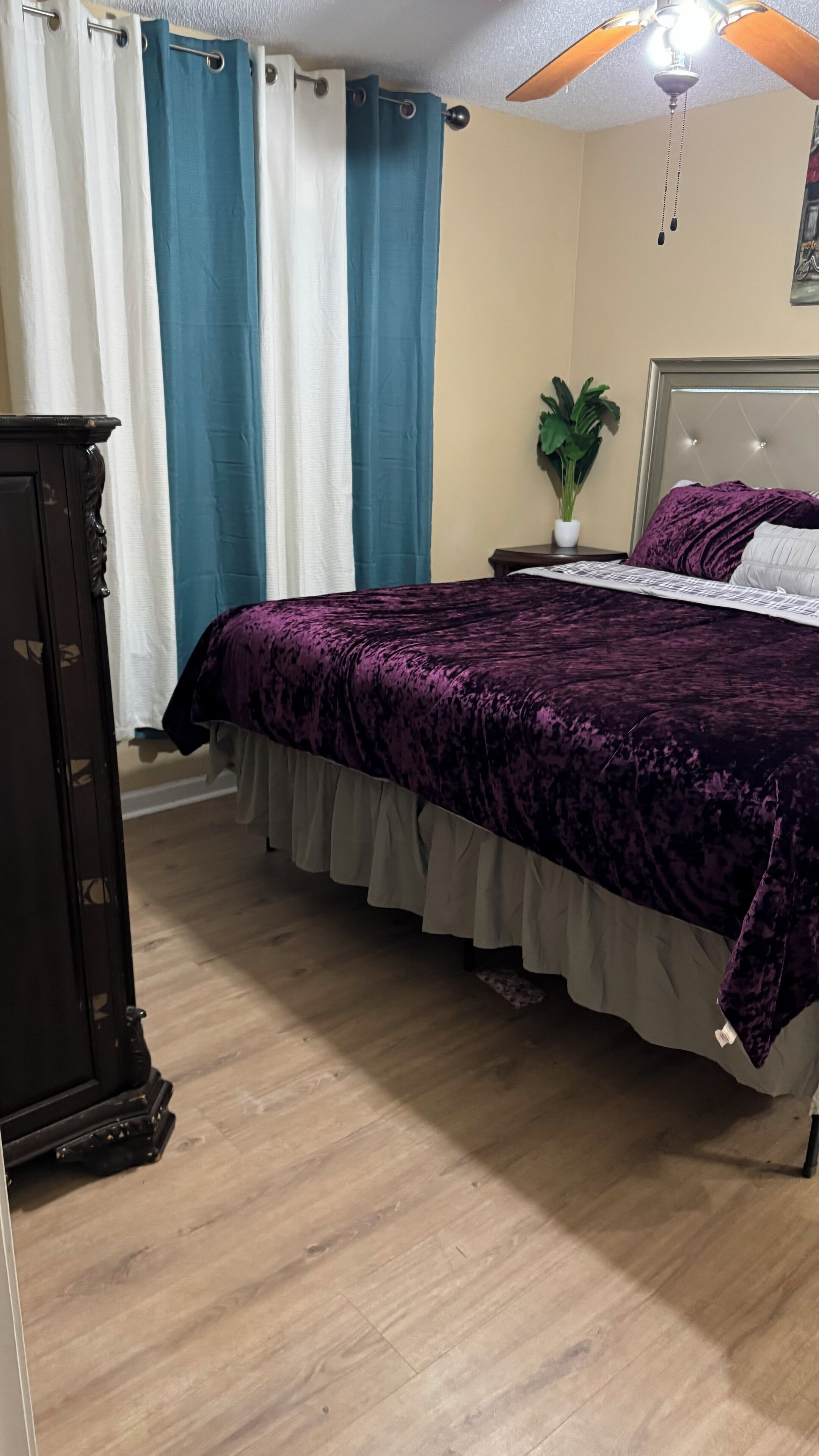 Un dormitorio con cama, cortinas, ventilador de techo, mesita de noche con planta y cómoda.