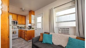 Un espacio habitable compacto con kitchenette, un sofá con cojines y un gran ventanal con cortinas.
