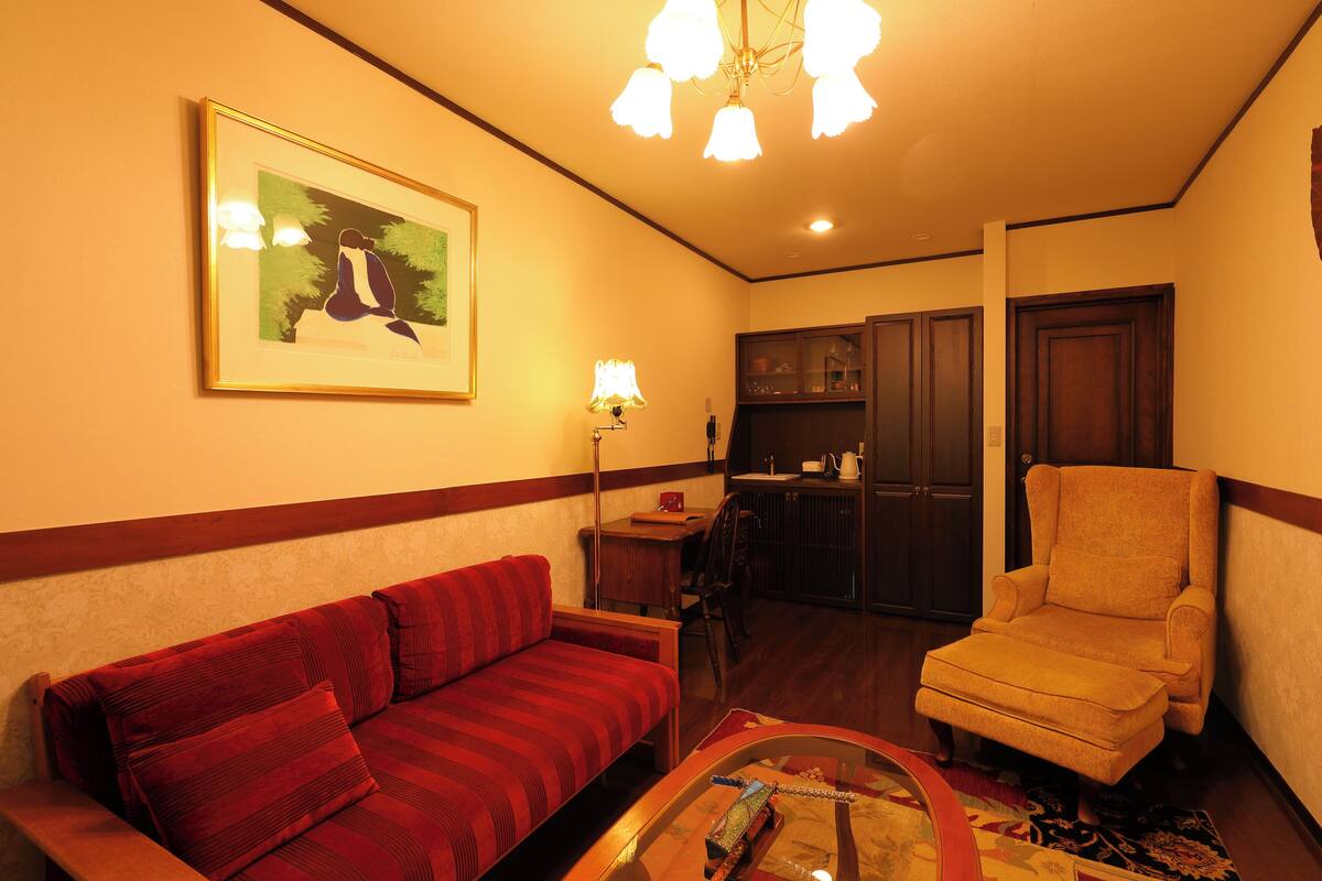 A living room with a red sofa, a yellow armchair, a coffee table, a framed picture on the wall, and a kitchen area in the background.
