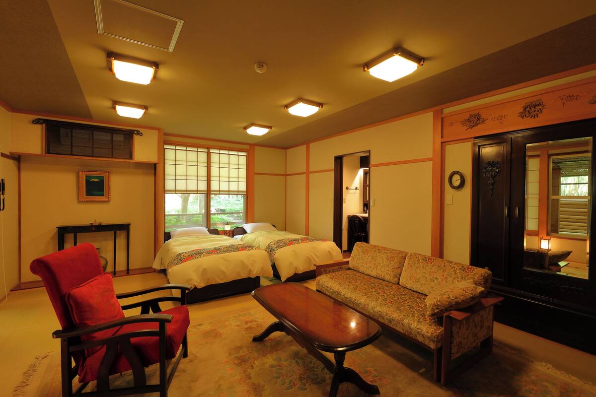 A traditional Japanese-style room with tatami flooring, a low wooden table, a red chair, a sofa, and two beds with tatami mats.