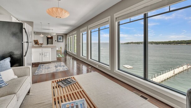 A spacious living area with large windows overlooking a body of water, a kitchen with white cabinets, and a modern pendant light. - Penthouse Suite, gorgeous West Lake Okoboji views, 1 BDRM above shopping center (Arnolds Park)