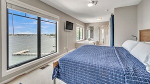 A bedroom with a large window overlooking a body of water, a bed with a blue quilt, a TV mounted on the wall, and a shower area. - Penthouse Suite, gorgeous West Lake Okoboji views, 1 BDRM above shopping center (Arnolds Park)