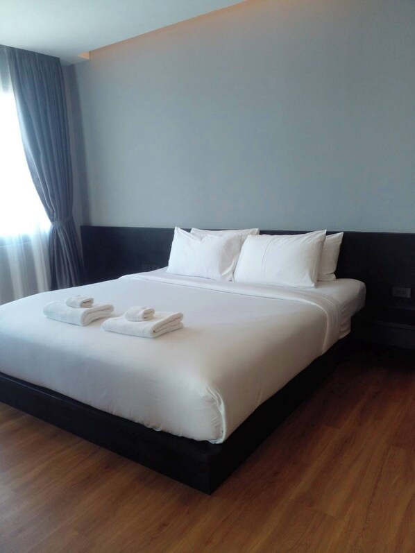 Uma cama bem arrumada com lençóis e almofadas brancos, piso de madeira e uma janela com cortinas.