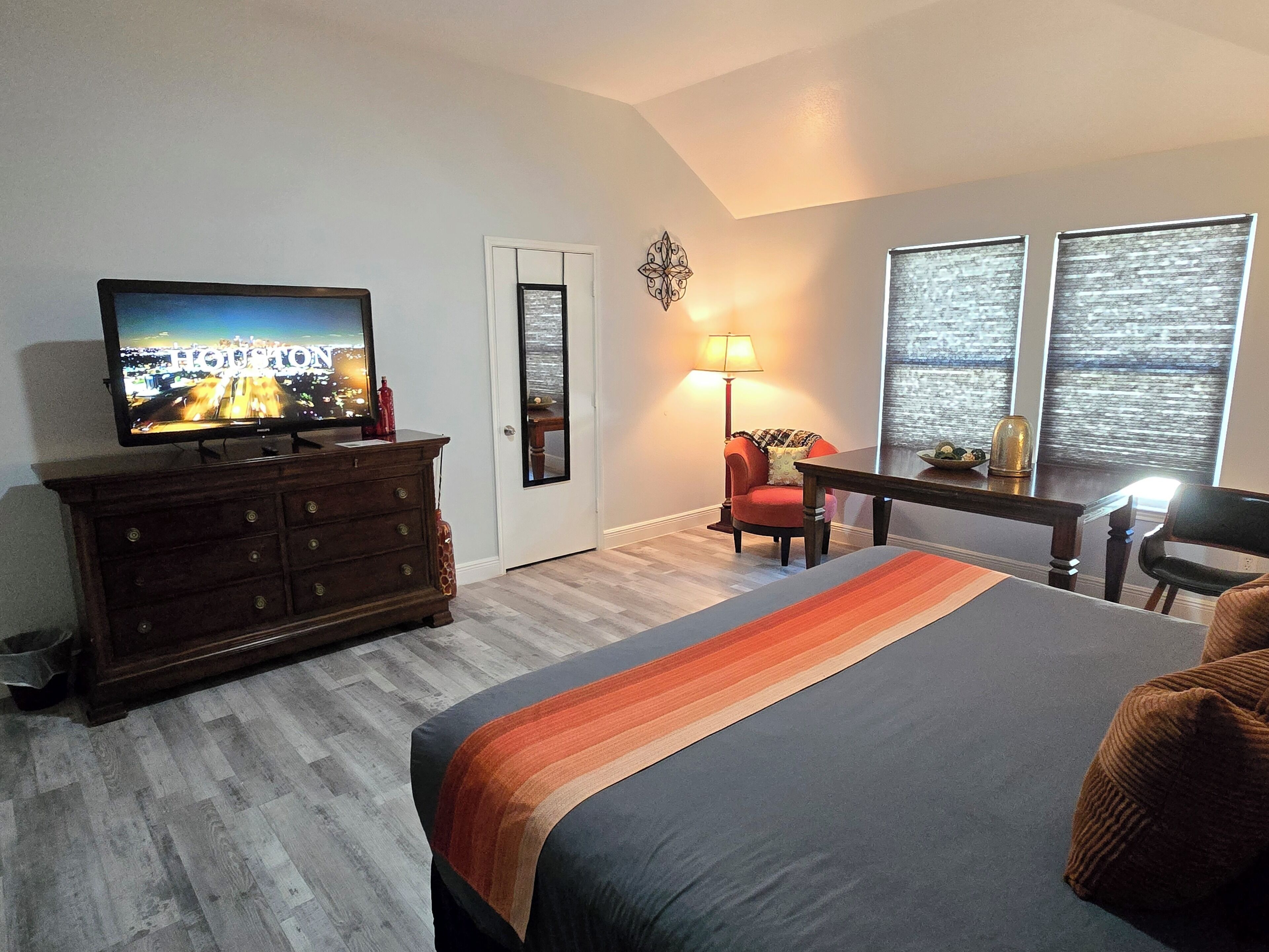A hotel room with a bed, a TV on a dresser, a desk with a chair, and two windows with blinds.