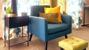 Une pièce avec un fauteuil bleu, une petite table d’appoint avec une lampe et un pouf jaune.