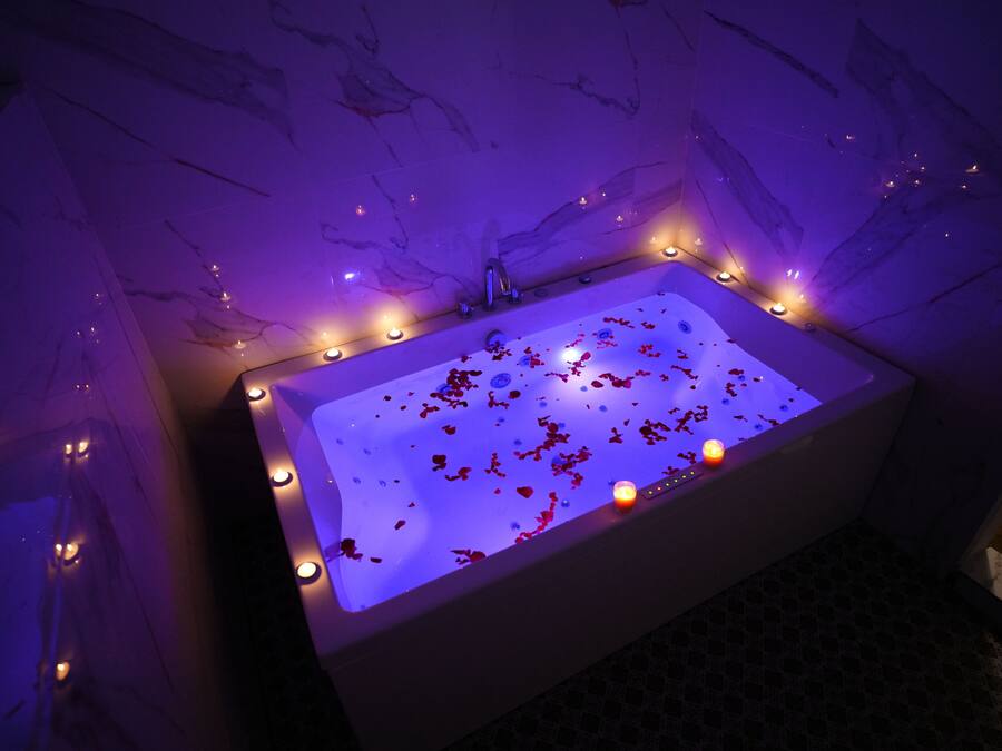 Una bañera rectangular con agua y pétalos de rosa, rodeada de luces empotradas en la pared.