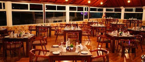A spacious restaurant with wooden tables and chairs, large windows, and a high ceiling.