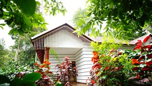 Een traditioneel huis met een schuin dak en een houten veranda, omringd door weelderige begroeiing en diverse planten.