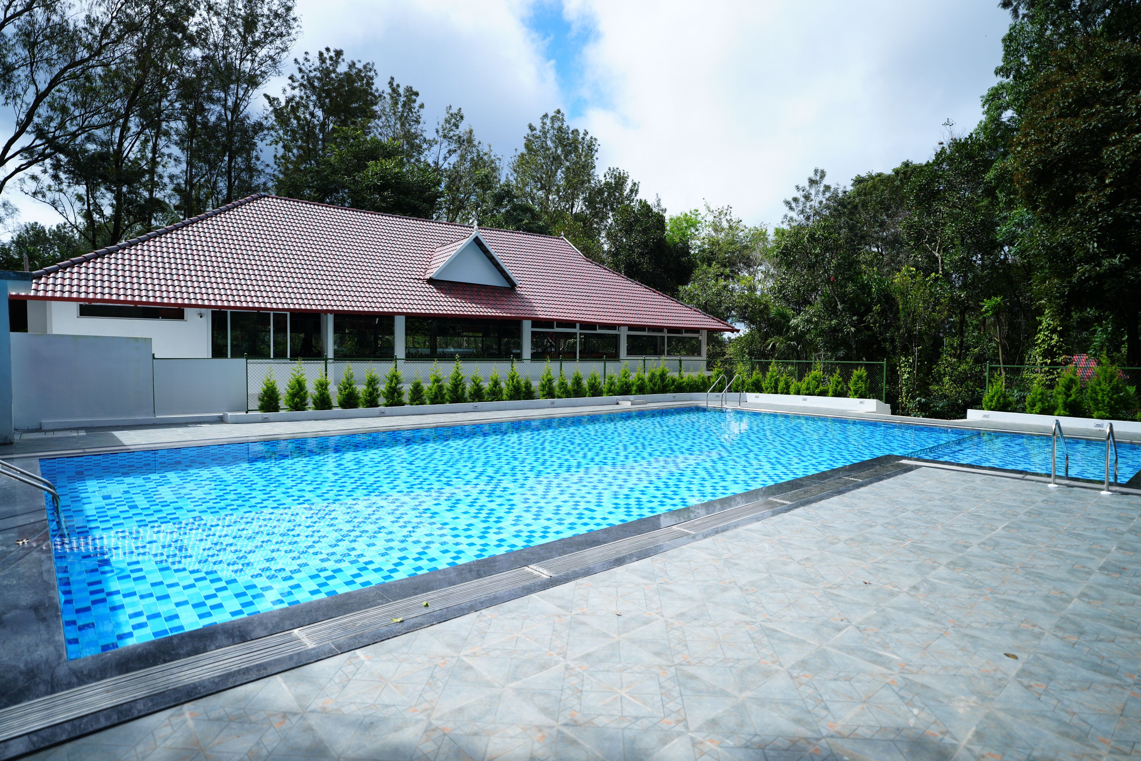 A swimming pool with a tiled deck, surrounded by trees and a building with a red roof.