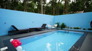 A swimming pool with lounge chairs and a small table, surrounded by a blue wall and trees.