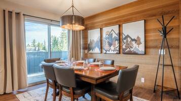 A dining area with a wooden table set for six, surrounded by chairs, with a view of trees through large windows.