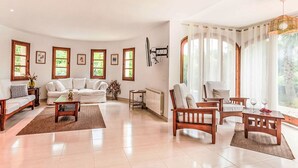 Un salon spacieux comprenant un canapé, des fauteuils, une table basse et une table à manger. De grandes fenêtres sont agrémentées de rideaux, et on y trouve également un radiateur et des tableaux encadrés accrochés au mur.