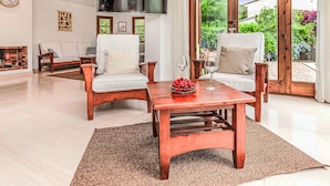 Un salon spacieux avec deux fauteuils en bois, une table basse sur laquelle se trouve un saladier rempli de fruits, et un téléviseur fixé au mur.