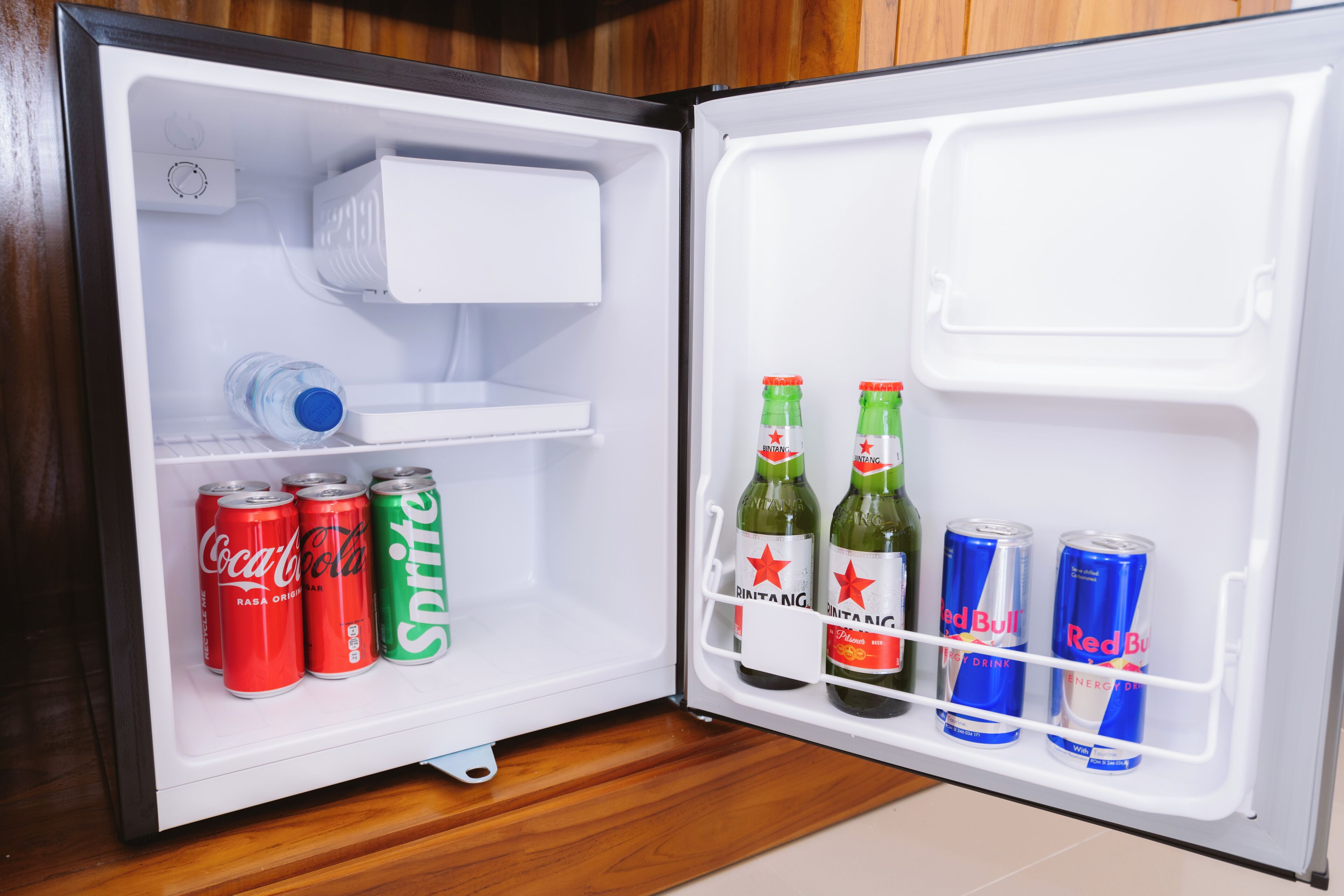 Un réfrigérateur ouvert, dont les étagères contiennent des bouteilles de Coca-Cola, de Sprite, de Red Bull et deux bouteilles de bière Brantang.