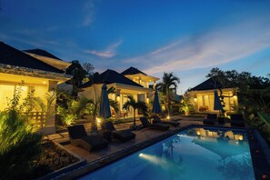 A poolside area with lounge chairs and umbrellas, surrounded by well-lit buildings and palm trees. - Kelingking Paradise Suites Villa (Penida Island)