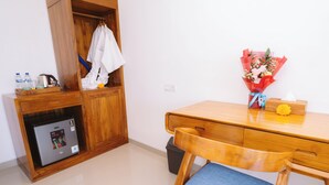 Ett hotellrum med ett skrivbord i trä, en stol med en blå kudde, ett litet kylskåp, en papperskorg, en garderob i trä och en bukett blommor på skrivbordet.