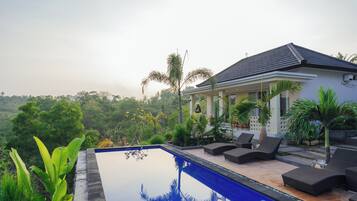 Una piscina con sillas de descanso, rodeada de exuberante vegetación y una casa con tejado de tejas.