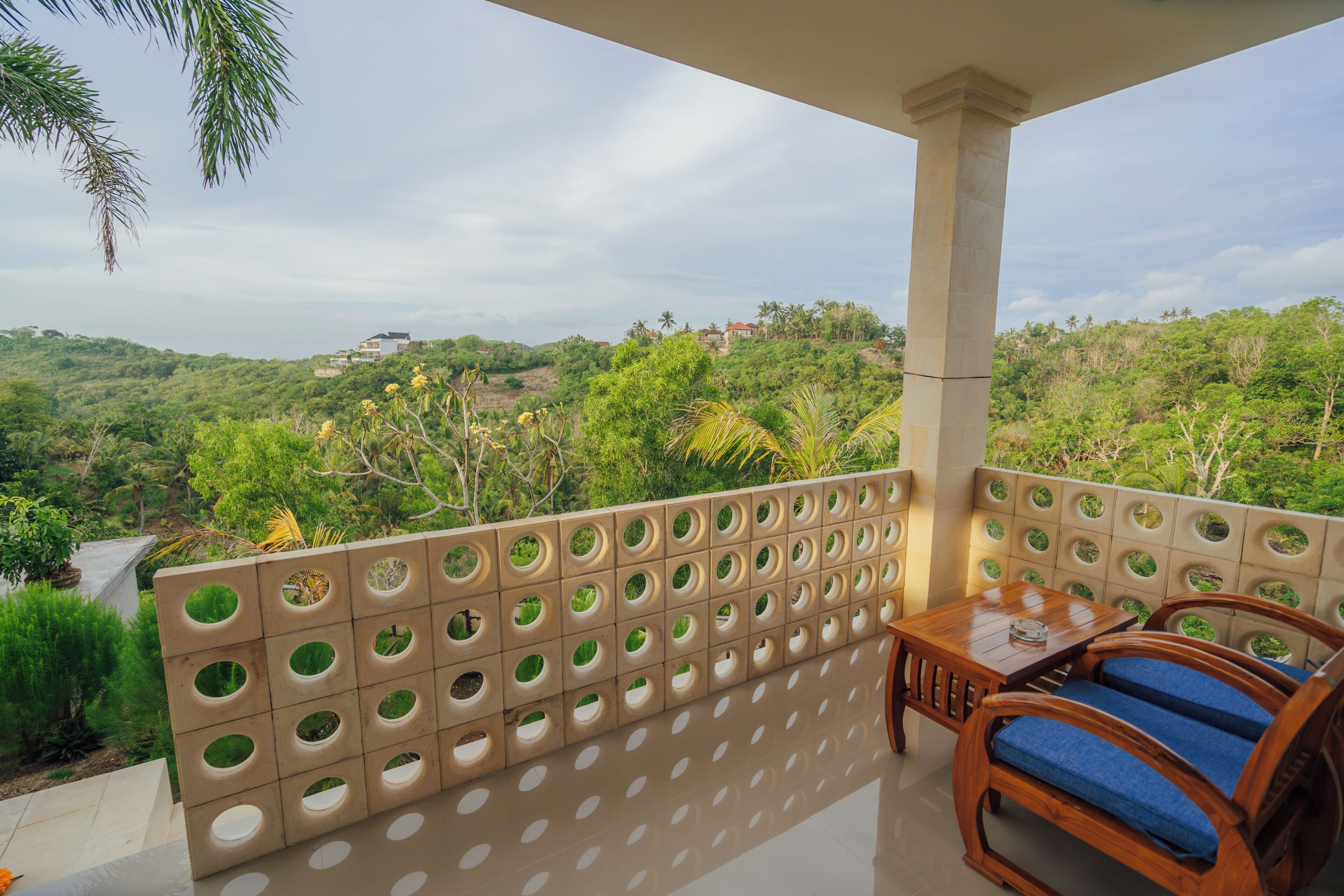 A balcony with a view of a lush landscape, a wooden table with chairs, and a perforated wall.