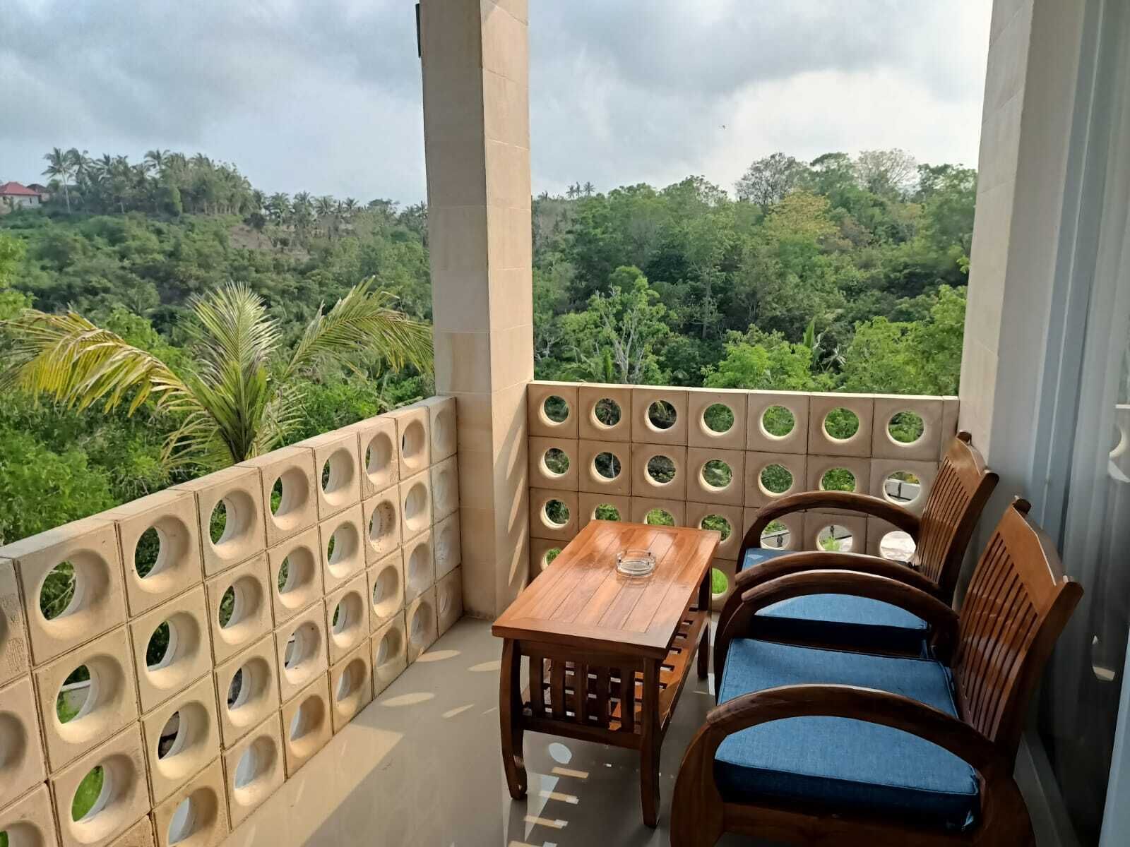 A balcony with a wooden table and two blue cushioned chairs, a beige perforated wall, and a view of a lush green landscape.