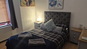 Une chambre à coucher avec une tête de lit capitonnée, une couvre-lit à carreaux, deux serviettes, une table de chevet avec une lampe et un tableau accroché au mur.