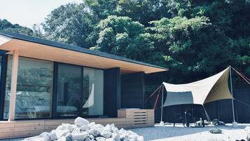 Una cabaña moderna, con paredes de vidrio y terraza de madera, rodeada por un paisaje natural y una tienda de campaña grande.