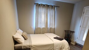 A bedroom with a bed, a bedside table, a chair, a waste bin, and a window with curtains.