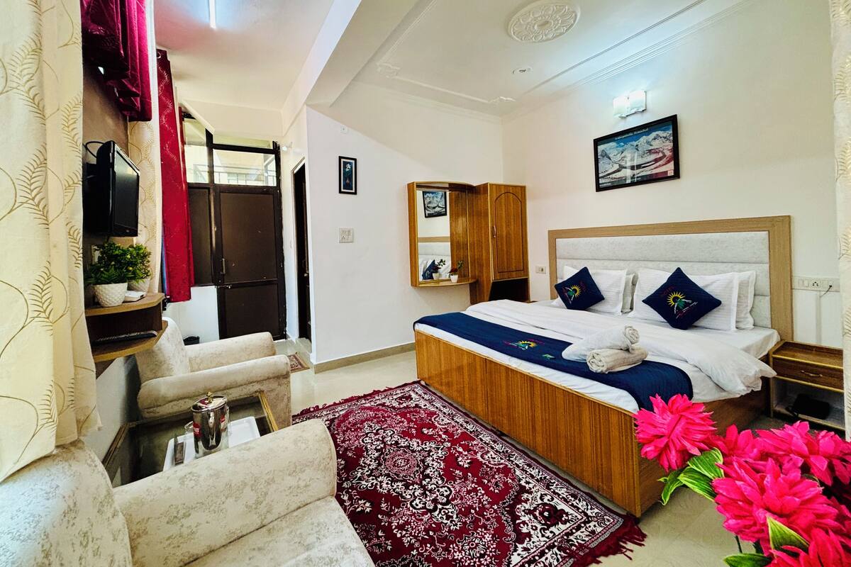A hotel room with a bed, a wooden wardrobe, a small table, a TV, and a red patterned rug.