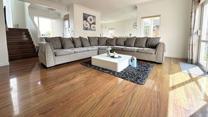 Ein geräumiges Wohnzimmer mit einer großen Eckcouch, einem weißen Couchtisch und einem schwarz-weißen Teppich. Im Hintergrund ist eine Treppe zu einer weiteren Ebene zu sehen.