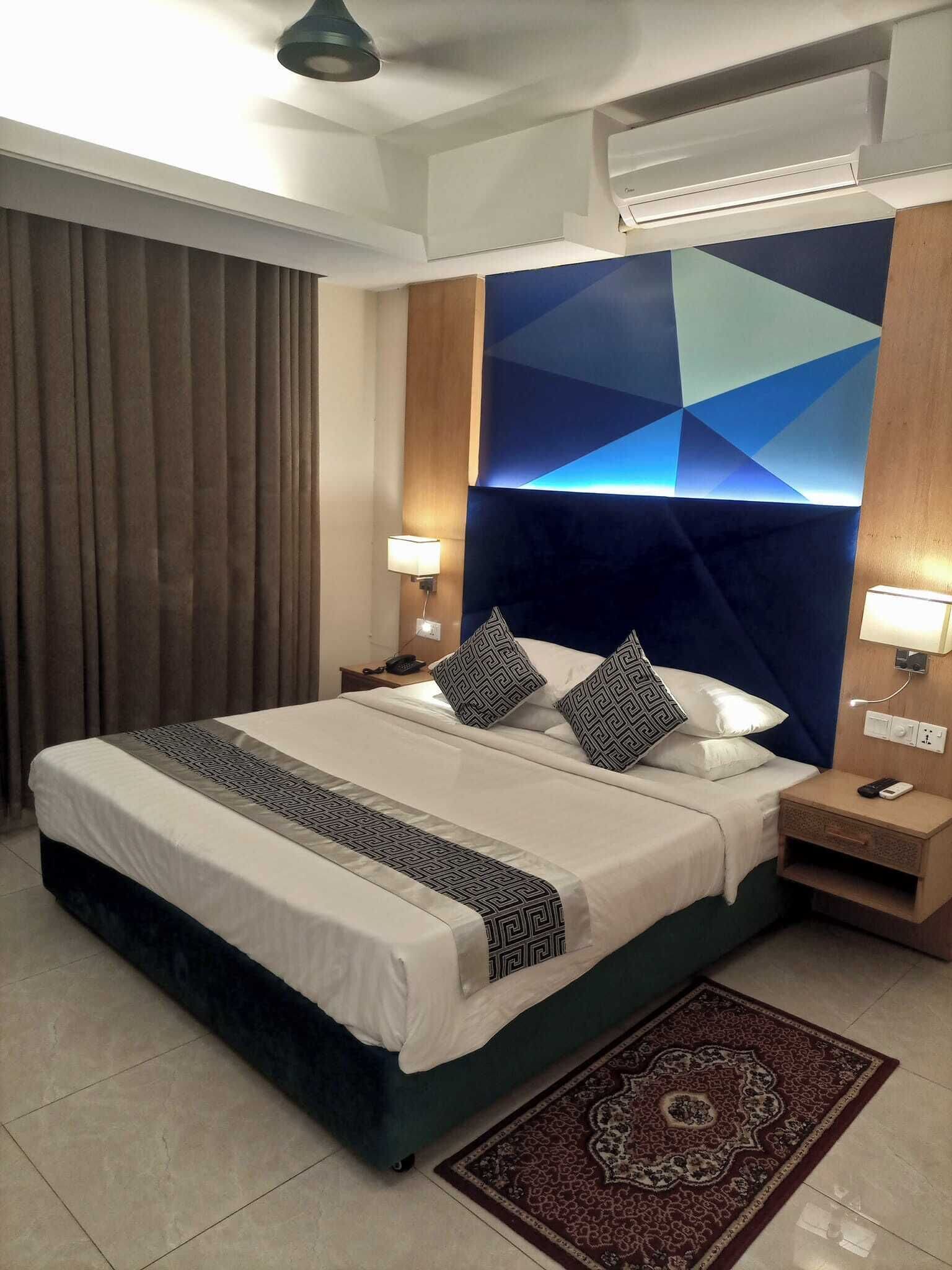 A hotel room with a large bed, two bedside tables, a wall-mounted air conditioner, and a patterned rug.