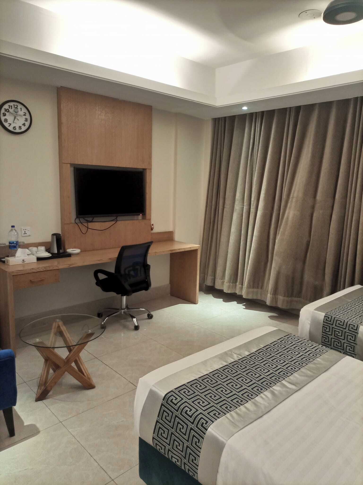 A hotel room with a desk, a television, two beds, a clock, and a window with curtains.