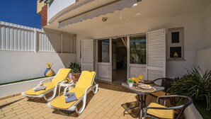 Een terras met gele loungestoelen en een tafel, een wit gebouw met luiken, en een klein raam met een lantaarn.
